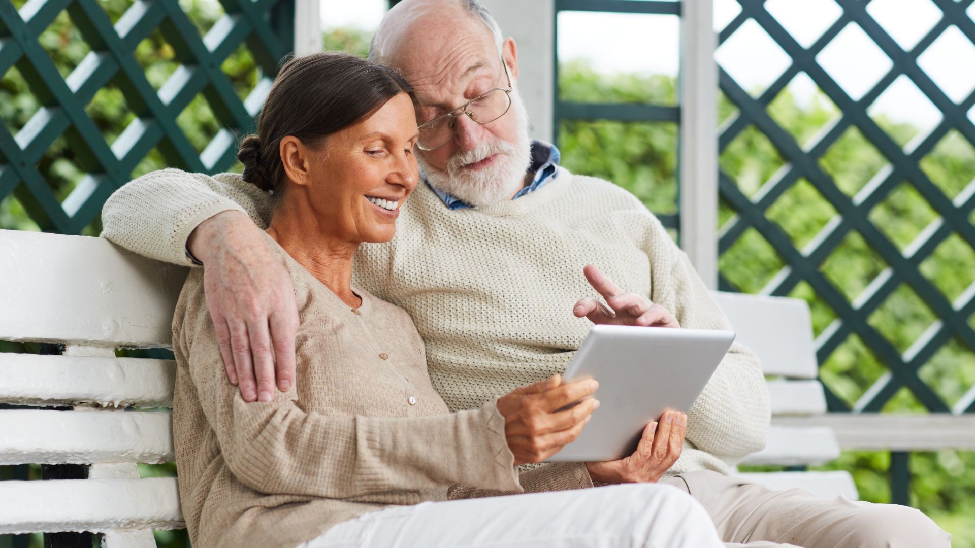 Couple âgé souriant avec tablette sur banc.
