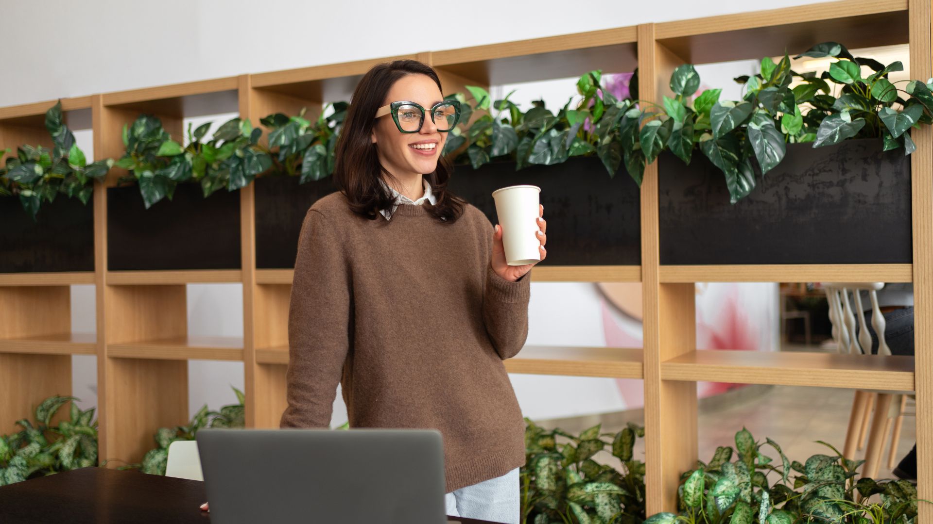 Femme souriante avec café devant ordinateur et plantes.