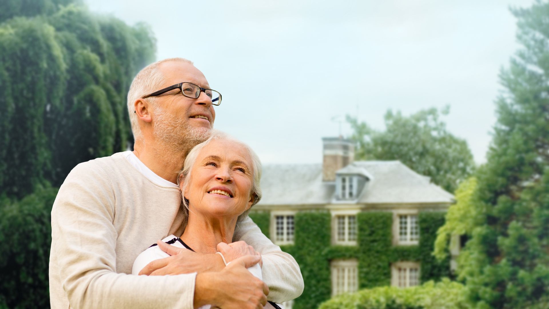 Couple âgé heureux devant maison verdoyante.